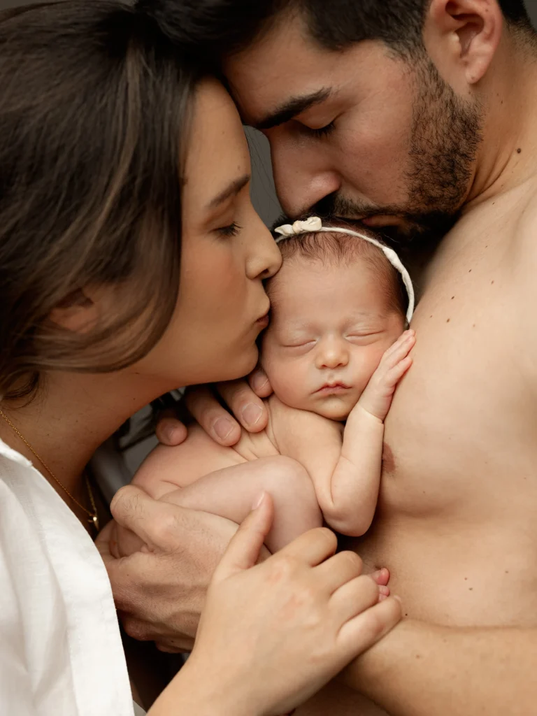 Um plano aproximado e íntimo de um casal a beijar suavemente a cabeça do seu bebé recém-nascido que dorme tranquilamente com uma fita na cabeça. A mãe está à esquerda de camisa branca e o pai à direita, abraçando o bebé contra o peito no estúdio fotográfico de Sofia de Melo.